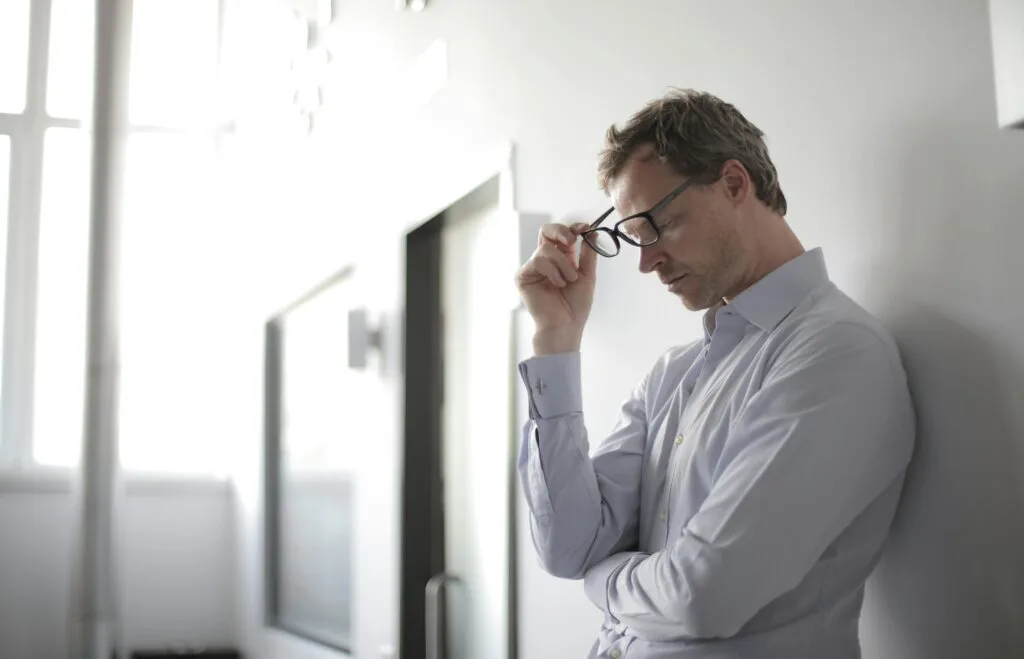 Thoughtful man in a bright room holding his glasses while leaning against a wall.