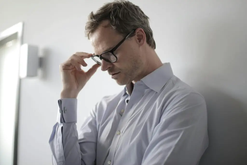 An adult man in a shirt and glasses appears stressed while leaning against a wall indoors.