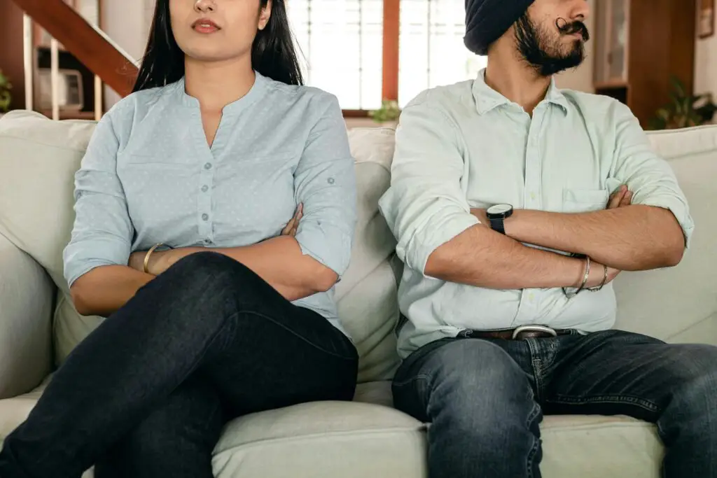 Crop faceless young ethnic couple in casual wear sitting on couch with arms crossed keeping silence after scandal at home